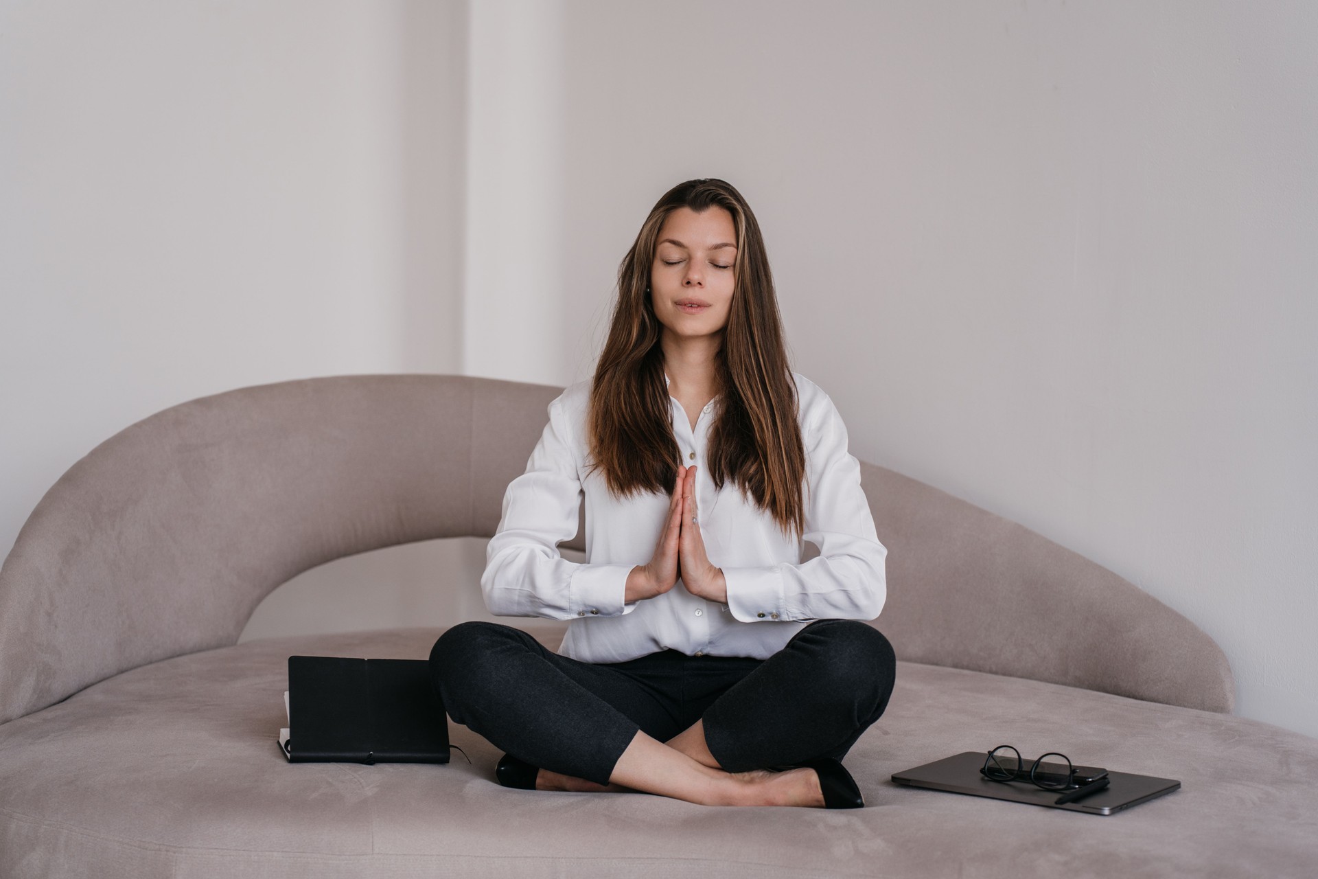 Mujer joven morena sobrecargada con camisa blanca, pantalones negros se sienta en un acogedor sofá en la postura de meditación piernas cruzadas con los ojos cerrados en casa. Bonita mujer de negocios relajante. Hembra con las manos juntas, reza.